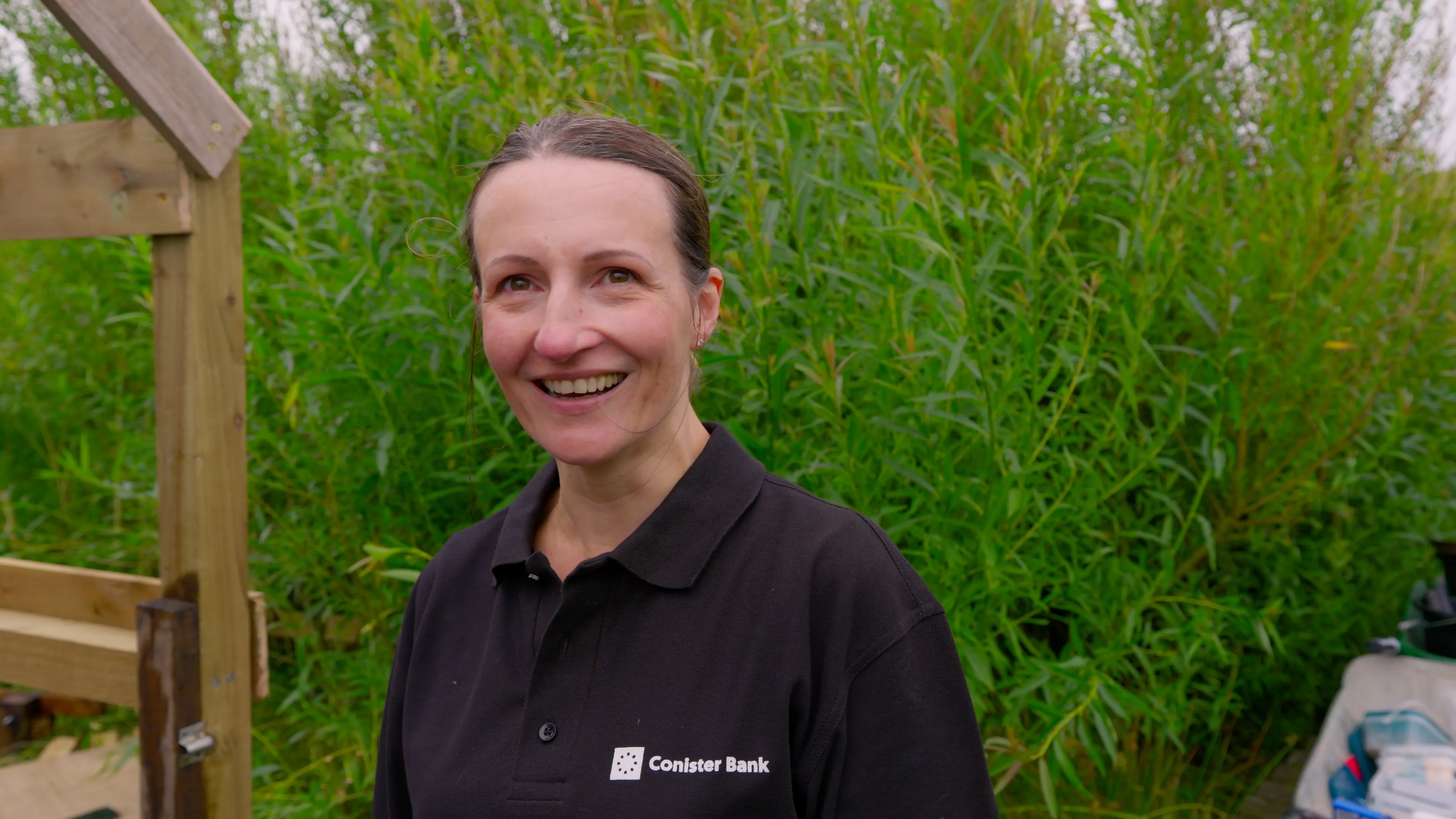 A smiling person in a black Conister Bank polo shirt stands outdoors in front of tall green bushes. The mood is cheerful and natural.